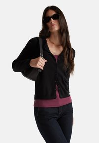 Zwarte cardigan met knopen, gedragen over een bordeaux top. Model houdt een zwarte tas vast, draagt donkere jeans en een zonnebril. Soepele texturen.