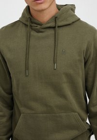 Olive green hoodie made of soft fabric, featuring a kangaroo pocket, drawstring hood, and subtle logo embroidery on the chest.