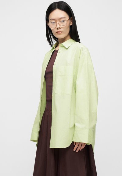 Femme aux longs cheveux noirs portant des lunettes transparentes, une chemise ample vert clair boutonnée sur une robe marron foncé, posant devant un fond uni.