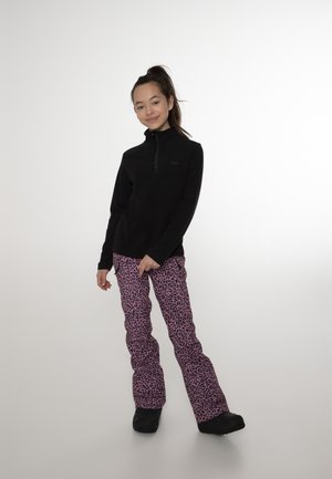 Jeune personne debout portant un haut noir à manches longues et un pantalon rose imprimé léopard avec des chaussures noires sur un fond uni.