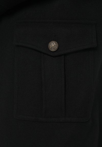 Abrigo negro de lana con un bolsillo de solapa y un botón metálico detallado. La textura parece suave y gruesa, adecuada para el clima frío.