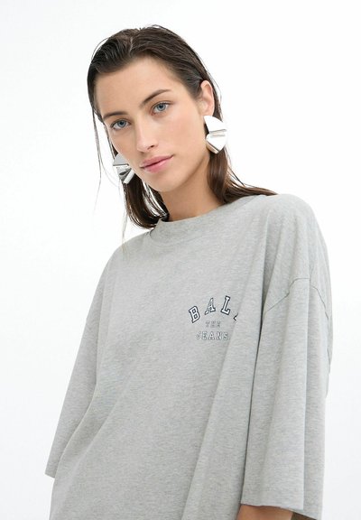 Šedé voľné tričko s nápismi "BAL" a "THE JEANS" v námornej modrej farbe. Modelka nosí veľké strieborné geometrické náušnice. Mäkký bavlnený materiál.