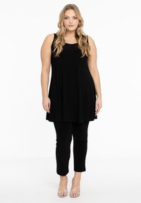 Schwarzes ärmelloses Tunika mit einem fließenden Design, kombiniert mit schwarzen Leggings. Das Modell trägt silberne Riemchensandalen. Ein einfacher und eleganter Look.