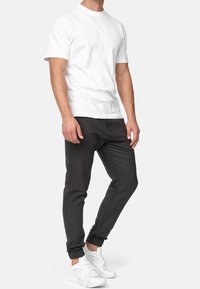 Mann trägt ein schlichtes weißes T-Shirt mit kurzen Ärmeln, dunkelgraue Jogginghosen mit elastischen Bündchen und weiße Sportschuhe, steht vor einem weißen Hintergrund.