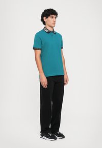 Ung mann med krøllete hår iført en teal piké med svart krage og kanter, svarte bukser og svart-hvite joggesko, stående på siden.