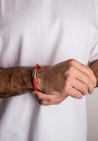 Raudonas pintas apyrankė su cilindrine metaline sagtimi, dėvima ant riešo su matomais plaukais, priešais vientisą baltą foną.