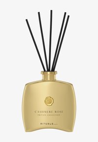 Reed diffuser med en gullfarget rektangulær flaske og et sirkulært emblem, med fem svarte rør som stikker opp fra toppen.
