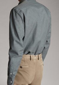 Camicia grigia chiara a scacchi con polsini abbottonati, abbinata a pantaloni beige con tasche posteriori e dettaglio in bottoni.