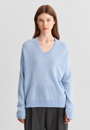 Jeune femme aux longs cheveux bruns portant un pull bleu clair à col en V et un pantalon ample gris foncé, debout devant un fond clair uni.