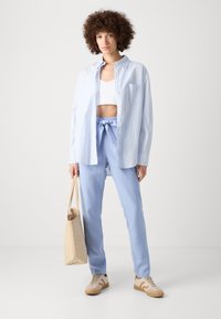 Blå og hvit stripete oversized skjorte, kort hvit topp, lyseblå bukser med knyteliv, beige tote-veske, og sneakers med brune detaljer.