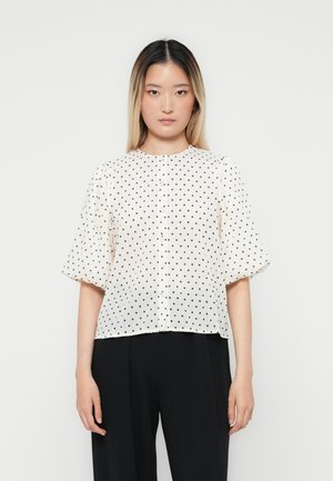 Blusa bianca a manica corta con pois neri, scollatura rotonda, dettagli a bottoni sul davanti e vestibilità ampia e fluida.