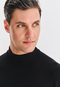 Czarny sweter z dzianiny z golfem i wysokim kołnierzem. Zdjęcie portretowe z miękkim oświetleniem podkreślającym rysy twarzy.