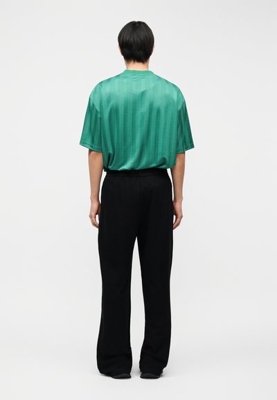 Άτομο που στέκεται πλάτη, φοράει πράσινο πουκάμισο με κοντά μανίκια και διακριτικές ρίγες και μαύρο παντελόνι με φαρδιά παντελόνια μπροστά σε απλό φόντο.