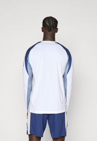 Langærmet træningsshirt i hvid med blå og lys grå detaljer. Har en rund halsudskæring og åndbart materiale. Kombinationen er med blå shorts.