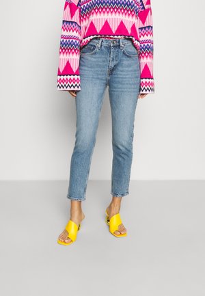 Person mit hoch taillierter blauer Jeans, einem rosa Pullover mit geometrischem Muster und gelben offenen Sandaletten mit Absatz, stehend auf hellgrauem Boden.