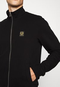 Svart zip-up sweatshirts med hög krage, med en liten guld-logotyp på bröstet och sidfickor, tillverkad i ett mjukt material.