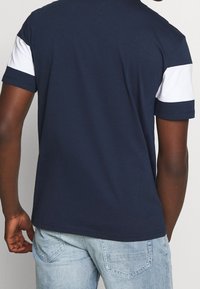 Osoba v námořnickém modrém tričku s bílými pruhy na rukávech a světle modrých džínách, pohled zezadu.