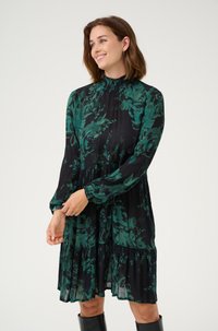 Musta mekko vihreällä kukkakuviolla, pitkät hihat, korkea kaula-aukko, smokkityyppinen yksityiskohta ja kerroksellinen röyhelöhelma. Silkkinen, kevyt kangas.