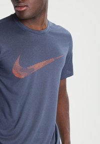 Man som har på sig en mörkblå Nike-tröja med orange strukturerad swoosh-logotyp, som tittar åt sidan mot en enkel ljus bakgrund.