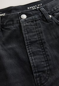 Gros plan sur un jean en denim noir avec fermeture par boutons et étiquette visible montrant "Baggy Fit 34 R".