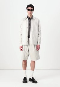 Lys grå skjorte med knapper og lomme, båret over en mørk ternet skjorte, parret med matchende shorts, hvide sokker og sorte sko.