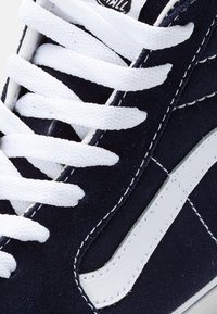 Námořnické plátěné tenisky s bílými tkaničkami a bočním pruhem. Vyznačují se bílým prošíváním a texturovanou tkaninou. Detailní záběr na design svršku obuvi.