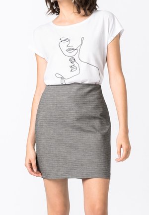 Femme portant un t-shirt blanc avec un dessin abstrait de visage en ligne et une mini-jupe grise texturée taille haute, debout devant un fond blanc.