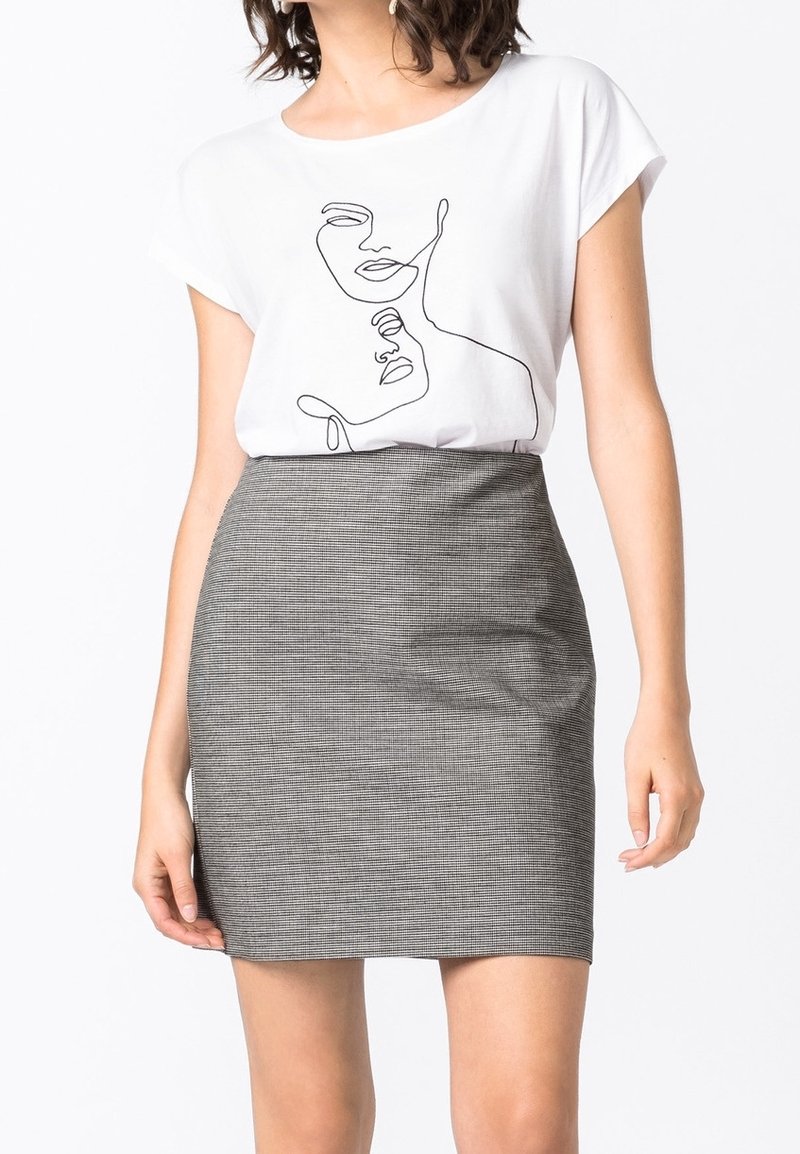 Femme portant un t-shirt blanc avec un dessin abstrait de visage en ligne et une mini-jupe grise texturée taille haute, debout devant un fond blanc.