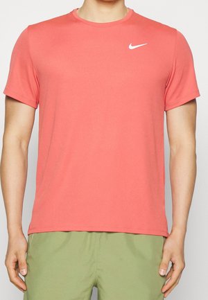 Homme portant un t-shirt Nike corail à manches courtes et un short vert clair, debout, bras détendus le long du corps, cadré au-dessus de la bouche.