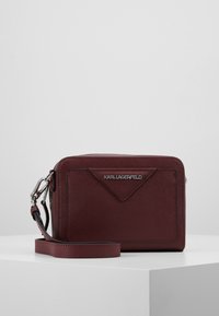 Vínově červená kožená crossbody taška se zipovým zapínáním, trojúhelníkovým předním designem, stříbrným logem a odnímatelným ramenním popruhem.