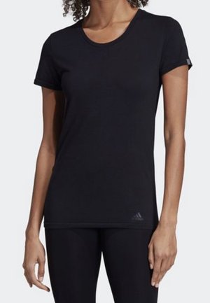 Frau trägt ein enganliegendes schwarzes Kurzarm-T-Shirt und schwarze Leggings, steht mit entspannten Armen an den Seiten vor einem schlichten Hintergrund.