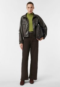 Kobieta w ciemno-brązowej skórzanej kurtce, zielonym golfie, ciemnych szerokich spodniach i brązowych botkach, trzymająca dużą, plecioną czarną torbę pod pachą.