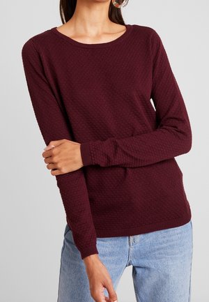 Vrouw draagt een getextureerde, donkerbordeauxrode trui met lange mouwen en een ronde hals, gecombineerd met lichtblauwe spijkerbroek, en houdt één arm over haar lichaam.