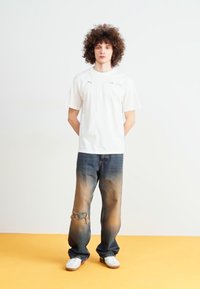 Camiseta blanca de manga corta con logo, combinada con jeans oscuros desgastados que presentan detalles de desvanecimiento y deshilachado. Zapatillas blancas completan el conjunto.