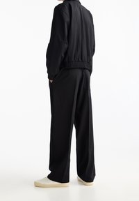 Schwarze Freizeitjacke mit elastischem Bund und Bündchen, kombiniert mit locker sitzenden schwarzen Hosen und weißen Turnschuhen. Glatter Stoff, einfaches Design.