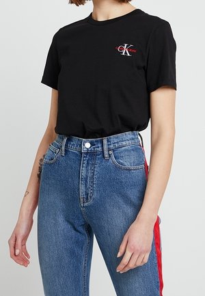 Schwarzes Calvin Klein T-Shirt mit kleinem Logo auf der Brust, kombiniert mit blauen, hoch taillierten Jeans mit rotem Streifen an der Seite.