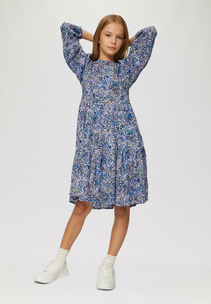 Kwiatowa sukienka w odcieniach niebieskiego, fioletowego i zielonego; długie bufiaste rękawy; falbaniasta spódnica; noszona z białymi trampkami i skarpetkami do kostek.