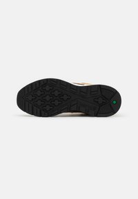 Semelle de chaussure en caoutchouc noir avec des motifs géométriques et un petit hexagone vert près du talon sur fond blanc.