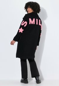 Cappotto in pile nero con testo rosa "SMILE" e stelle, maniche lunghe e silhouette rilassata. Indossato sopra pantaloni neri e stivali chunky.