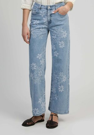 Jean large bleu clair avec des motifs floraux blancs porté avec des sandales noires à brides et un chemisier blanc à bordure en dentelle.