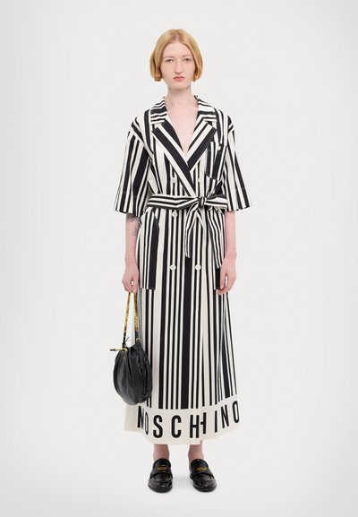 Melna un balta svītraina garā toga ar jostām viduklī, kurai ir lielie pogas un vārds "MOSCHINO" apakšmalā. Modelis tur melnu rokassomu.