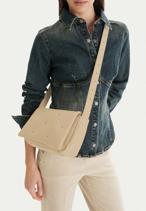 Mujer que lleva una chaqueta vaquera oscura y pantalones beige con un bolso bandolera beige que presenta pequeñas tachuelas metálicas en la solapa.