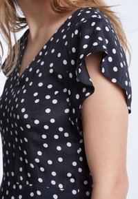 Robe noire à pois blancs, décolleté en V, manches courtes à volants et texture de tissu lisse. Les détails mettent l'accent sur les bords des manches et le motif.