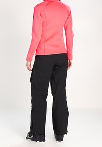 Haut à manches longues de couleur rose vif, associé à un pantalon noir et imperméable. Les pantalons disposent de zips aux chevilles et d'un discret détail de logo.