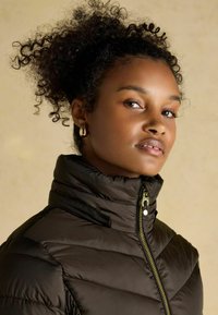 Hnedý hrubý kabát s vysokým golierom, diagonálnym prešívaním a zlatým zipsom. Modelka s kučeravými vlasmi pózuje pred neutrálnym pozadím.