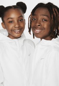 Dos niños que llevan chaquetas con capucha blancas de Nike con logotipos plateados, mostrando un tejido ligero y un diseño simple.