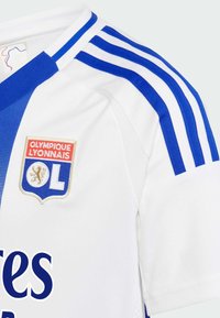 Bílý sportovní dres s modrými pruhy na ramenech, modrým výstřihem a logem Olympique Lyonnais na hrudi.