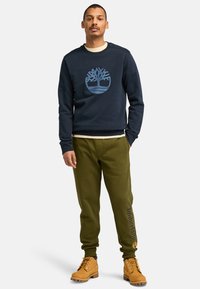Marineblauer Sweatshirt mit blauem Baumlogo, grüne Jogginghosen mit Timberland-Schriftzug und beigefarbene Wanderschuhe. Lässiges Outfit mit lockerer Passform.