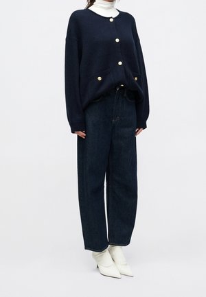 Kvinde iført sort knapstrikket cardigan, hvid højhalset trøje, mørke vide jeans og hvide spidse ankelstøvler mod en ensfarvet baggrund.