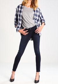Dark blue skinny jeans, grey t-shirt, and a black and white plaid shirt unbuttoned. High-heeled black shoes. Smooth textures throughout.